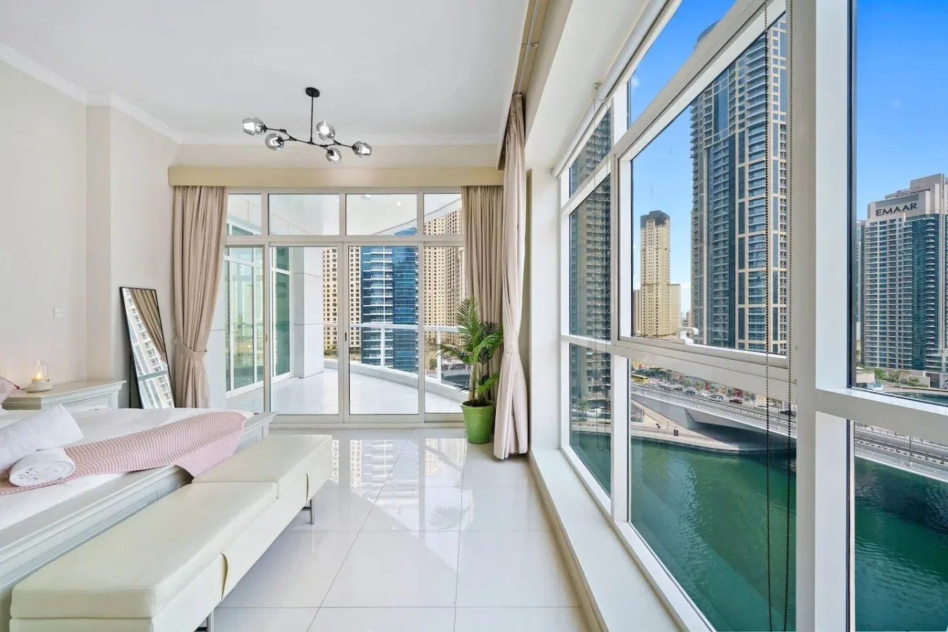 A bright bedroom with a bed, large windows, and views of high-rise buildings and water.