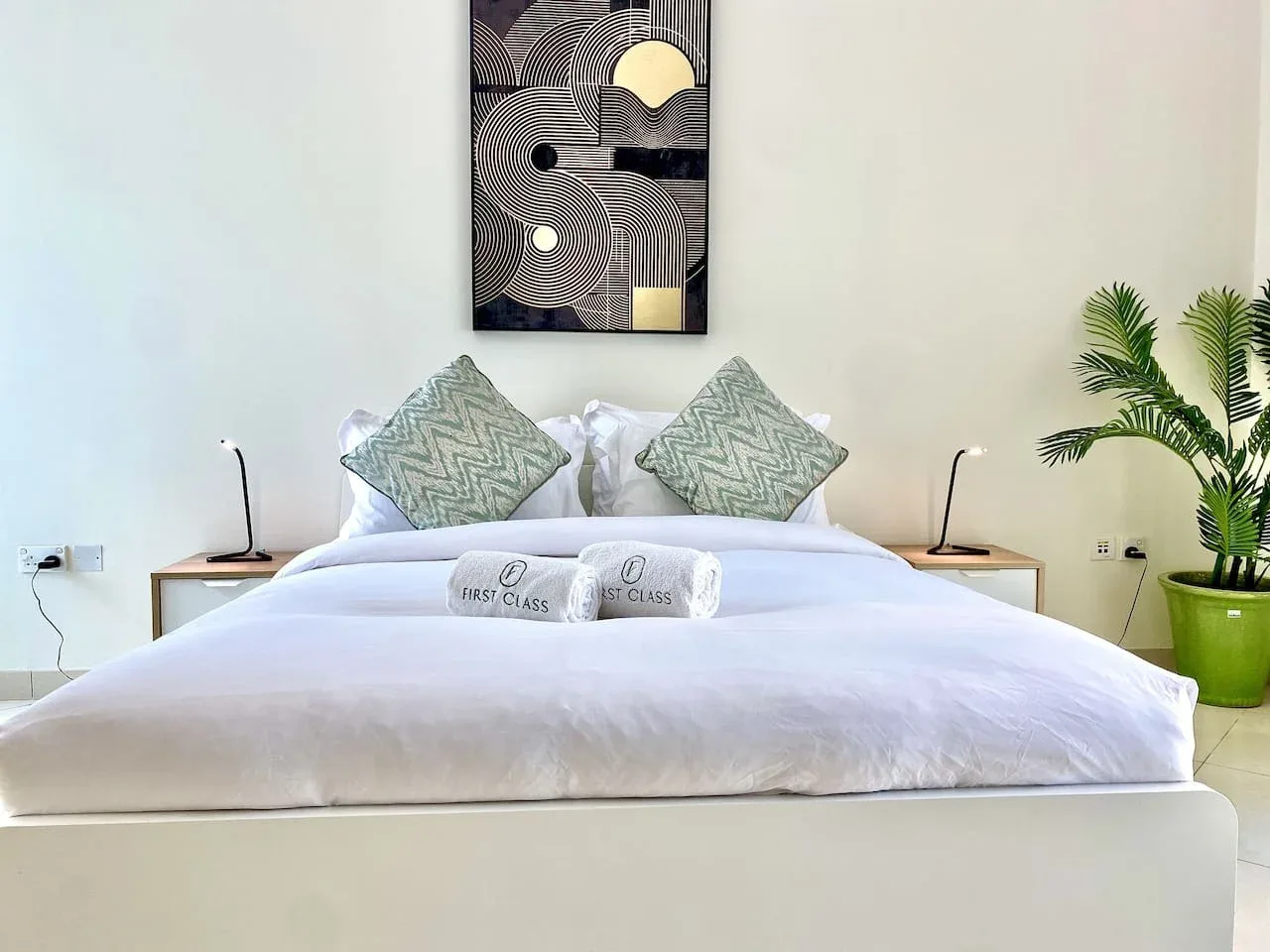 A bright bedroom with a neatly made bed and framed geometric artwork above it.