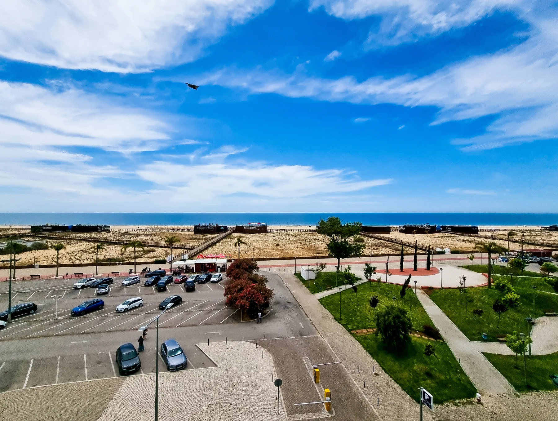 A coastal view overlooks a sandy beach, parking area, and green park.