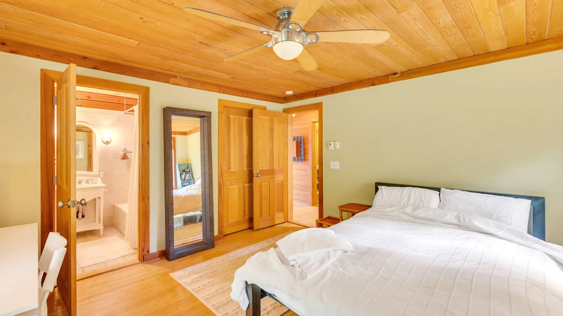 Dormitorio con una cama blanca, techo de madera y una puerta abierta hacia el baño.
