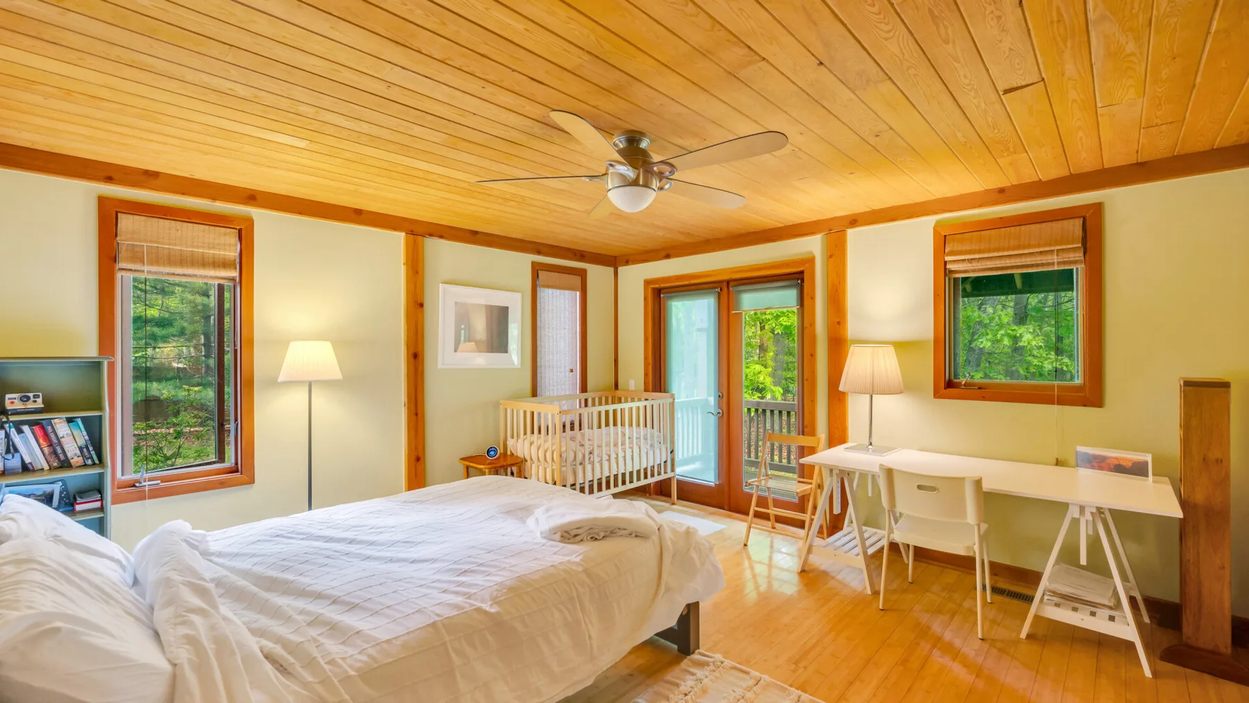 Un dormitorio luminoso con una cama grande, una cuna y un escritorio junto a las ventanas.
