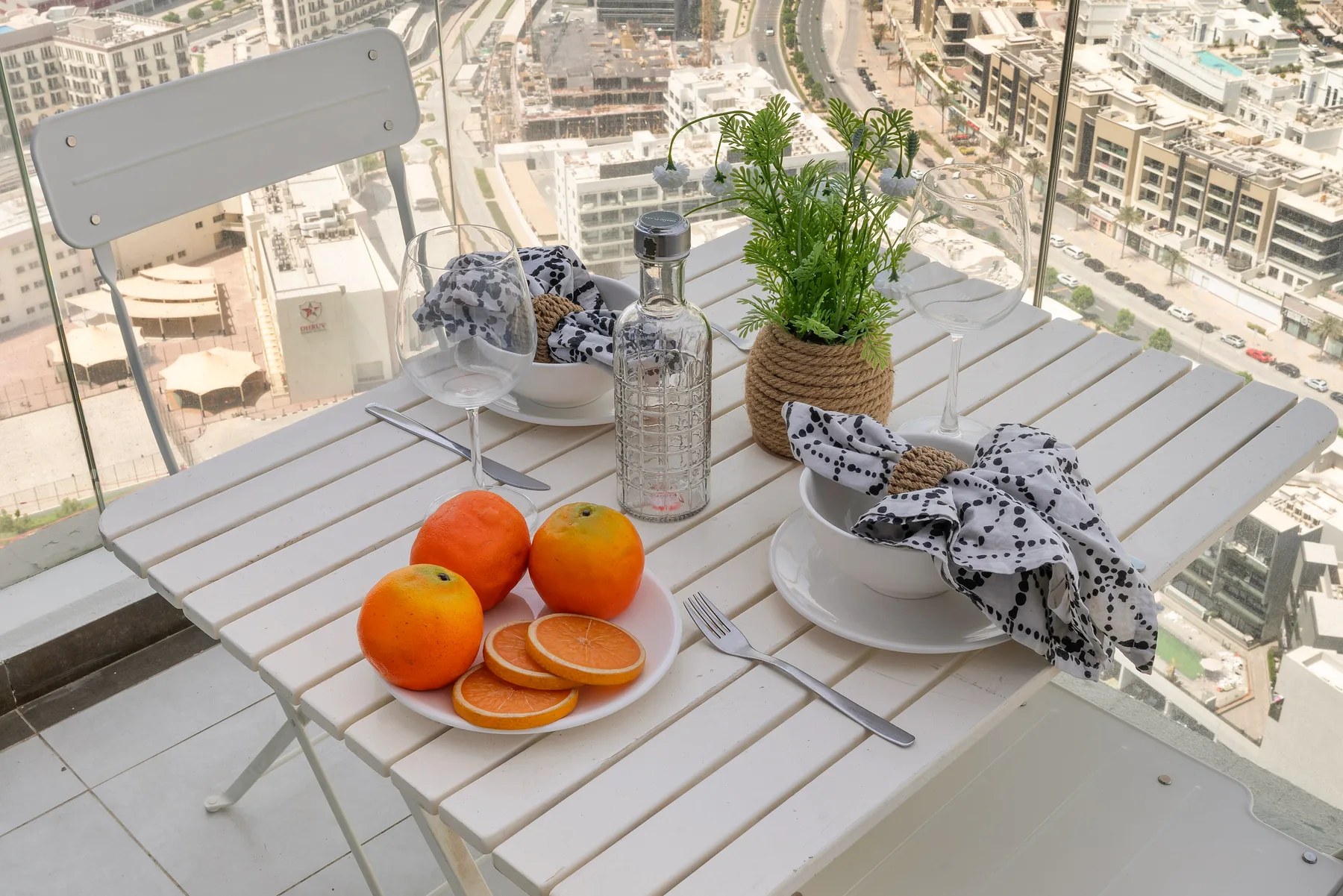 Una mesa de comedor en un balcón tiene naranjas, platos y una planta en maceta.