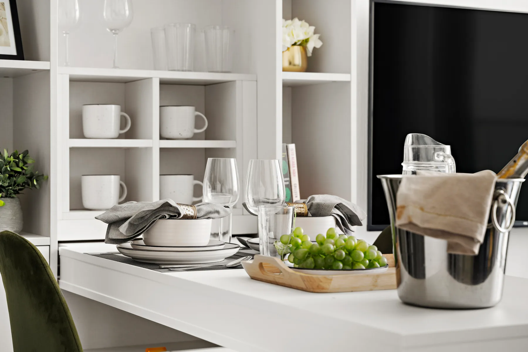 Una mesa de comedor con platos, copas y un recipiente con uvas frente a estantes abiertos.