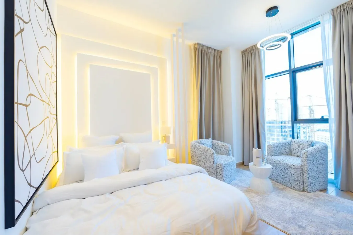 A bright bedroom with a white bed, upholstered seating, and a large window.