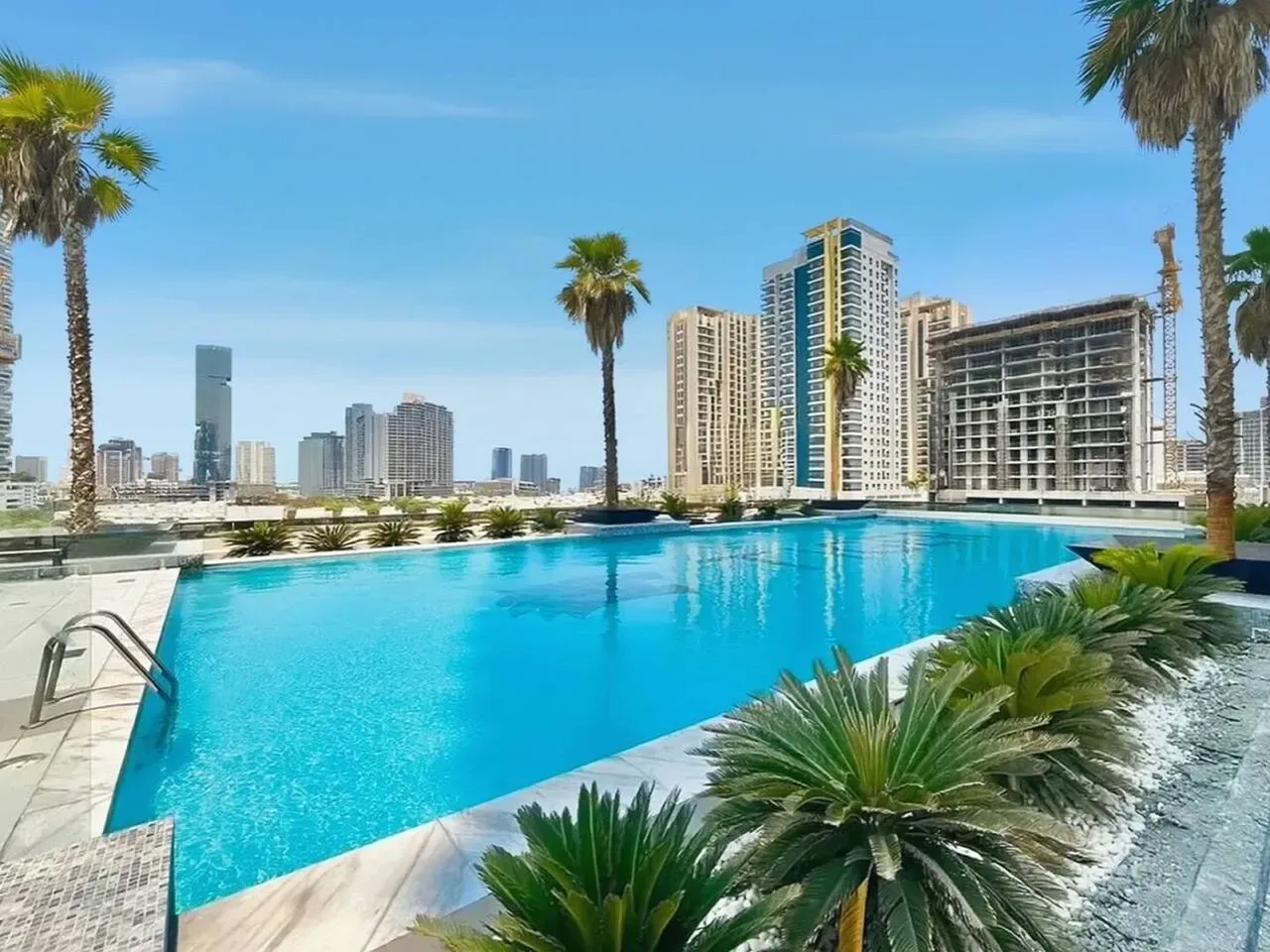 A rooftop swimming pool overlooks a cluster of high-rise buildings, with palm trees along the edge.