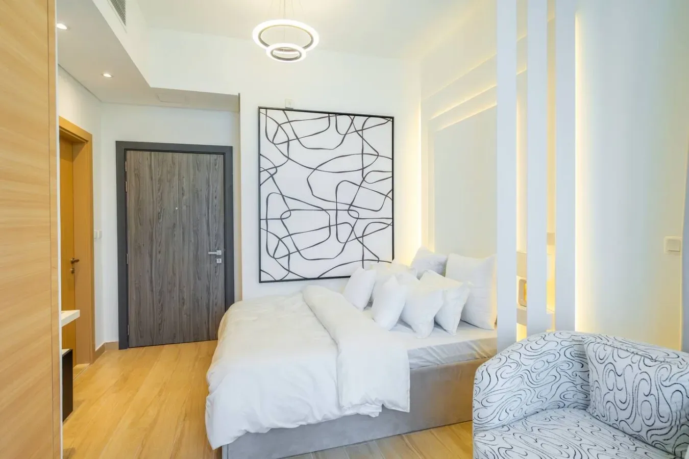 A bright bedroom with a white bed, abstract wall art, and a patterned armchair.