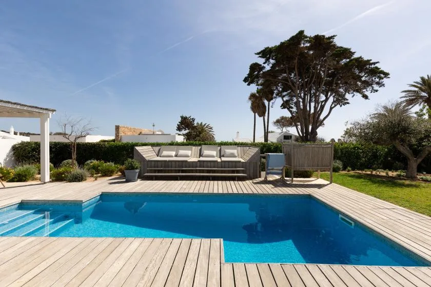 An outdoor swimming pool sits beside a wooden deck and cushioned seating.
