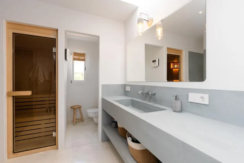 A bright bathroom with a long sink counter and a wooden sauna door.