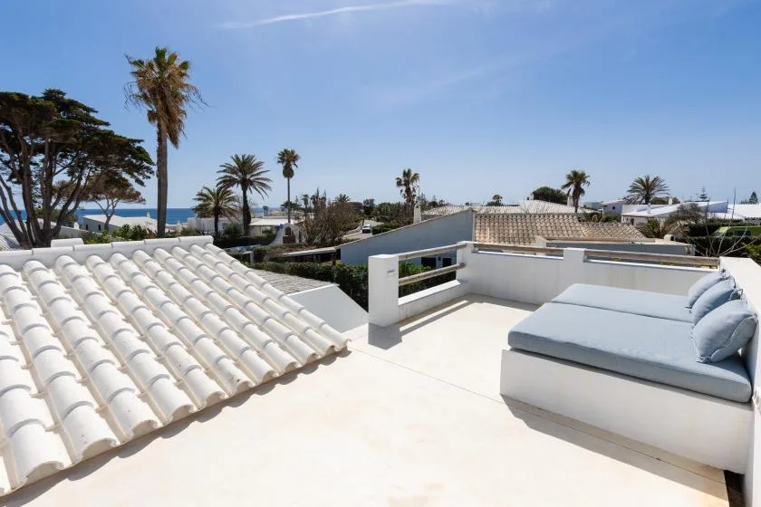 A sunny rooftop terrace with cushioned seating and views of palm trees and the sea.