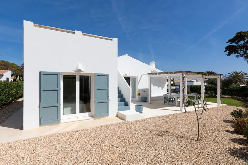 White stucco house with blue shutters, a gravel yard, and a shaded outdoor seating area.