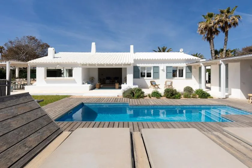 A white house faces a swimming pool, with lounge chairs on the surrounding deck.