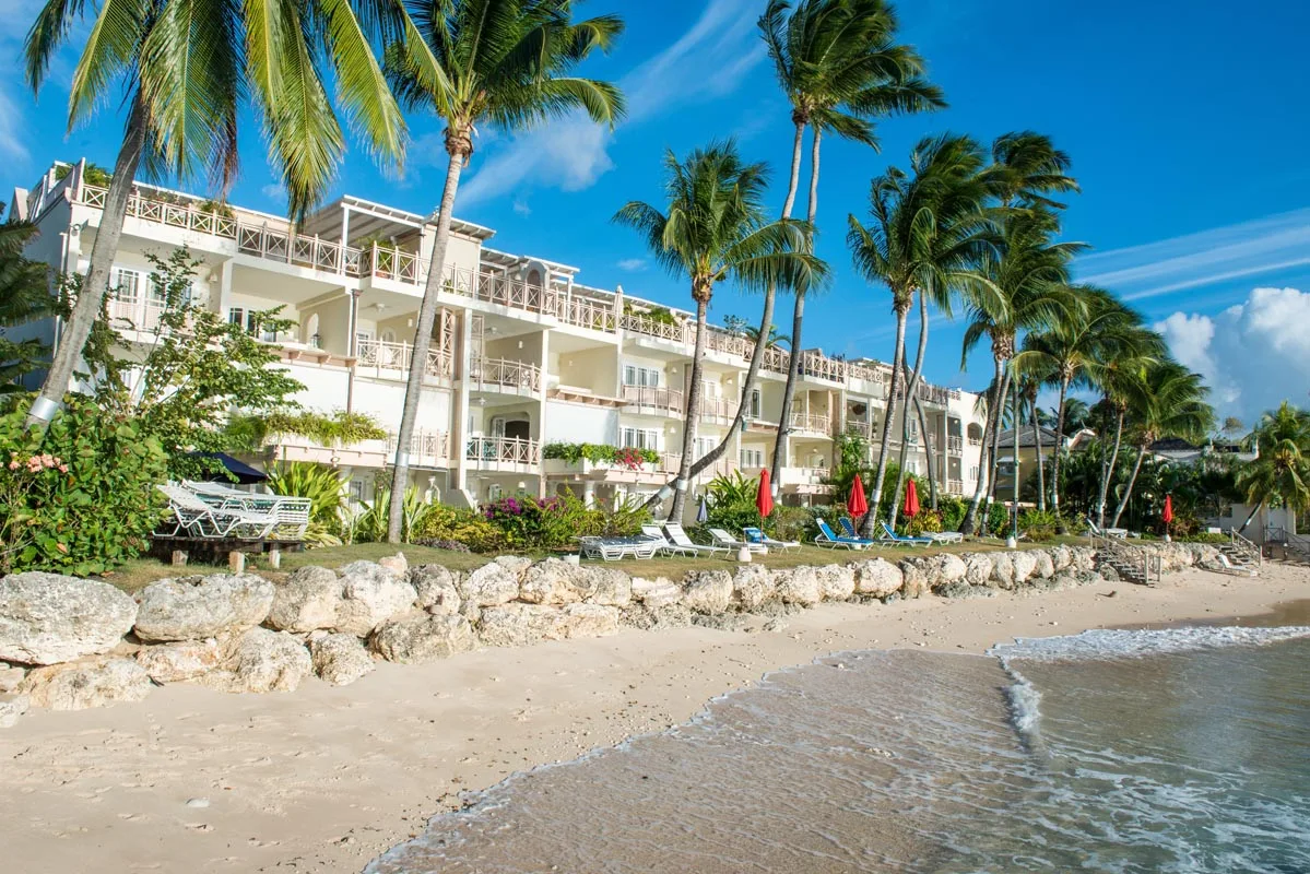 A beachfront resort building stands behind a sandy shore lined with palm trees.