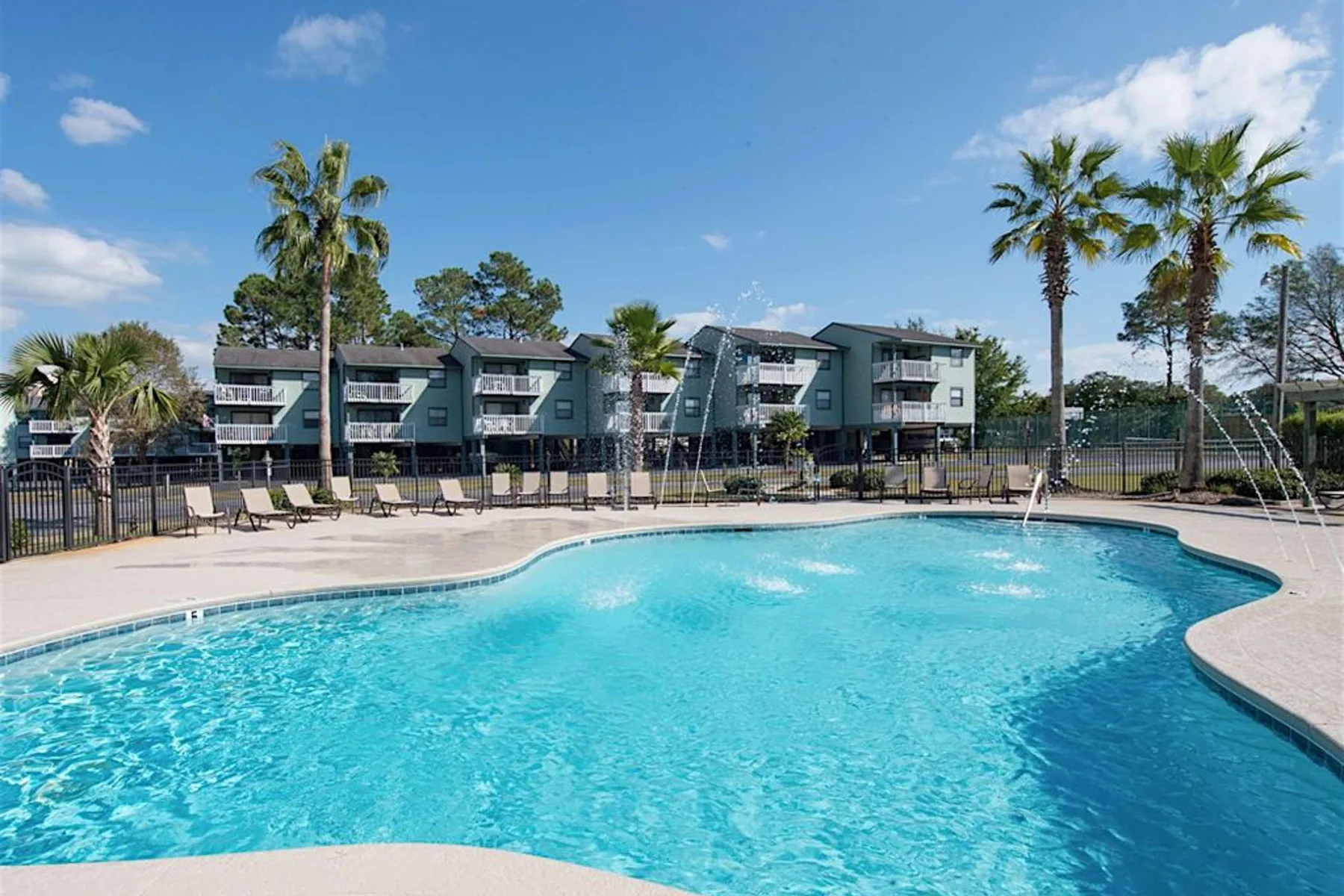 Una piscina junto a una terraza cercada, con edificios de apartamentos y palmeras al fondo.