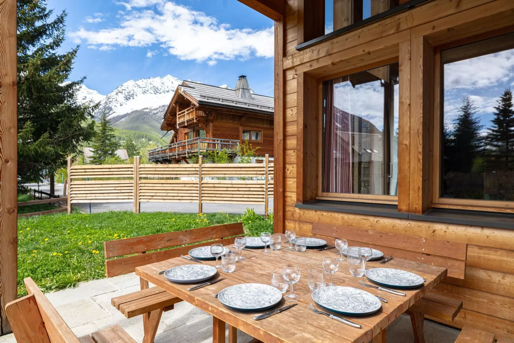 A wooden outdoor dining area overlooks a grassy yard and mountain scenery.