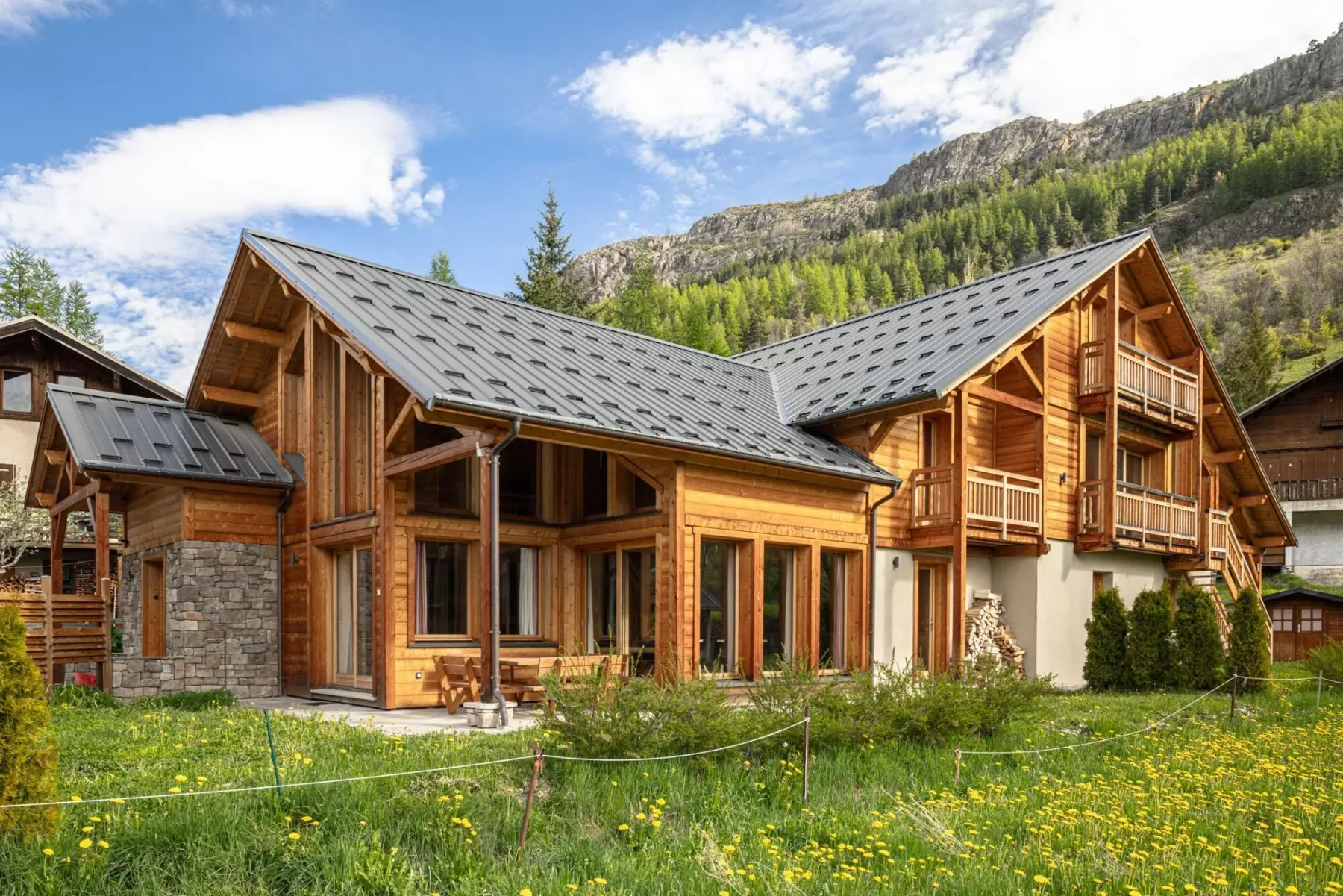 A wooden mountain lodge stands beside a grassy meadow under a blue sky.