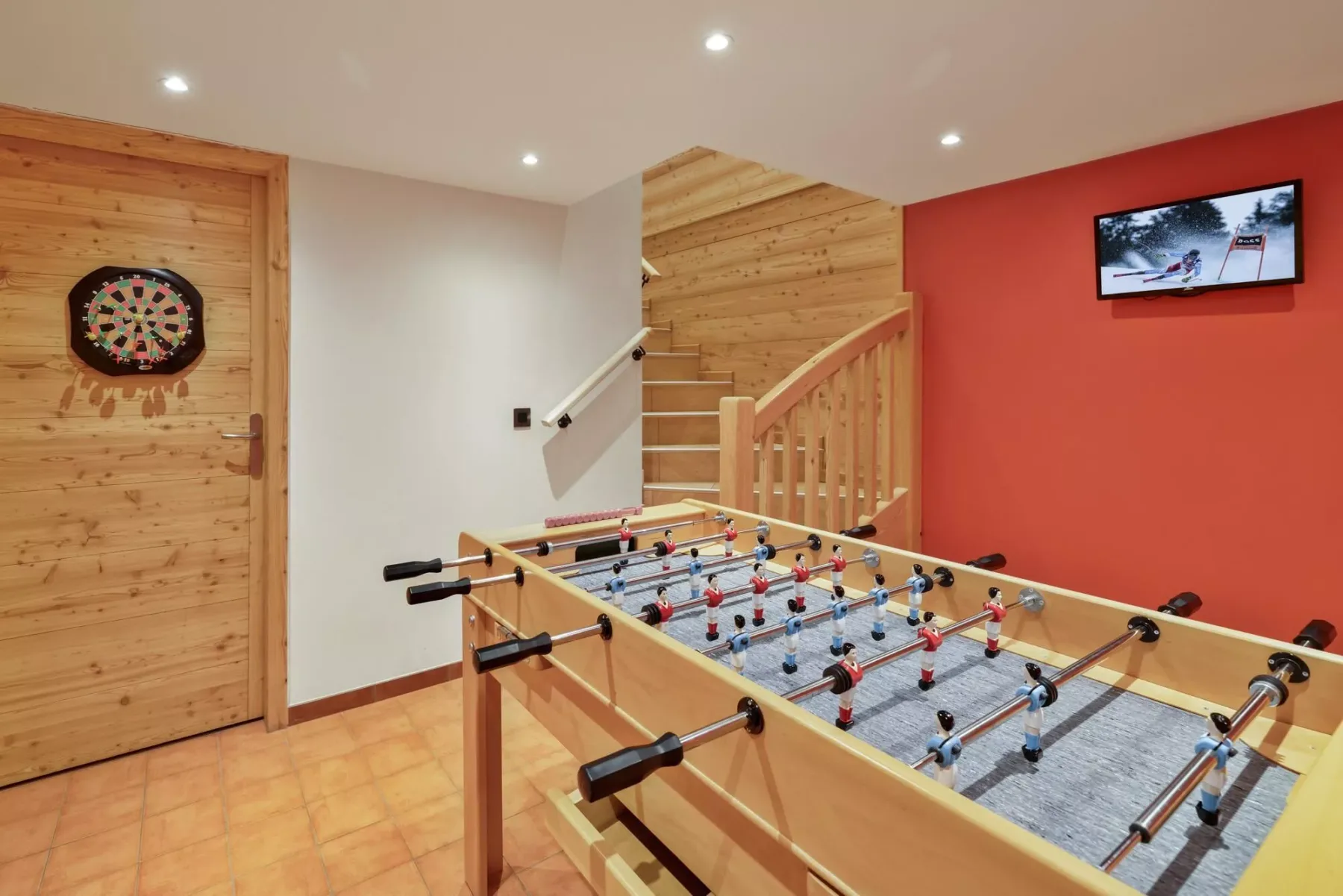 A recreation room with a foosball table, dartboard, and wall-mounted screen.