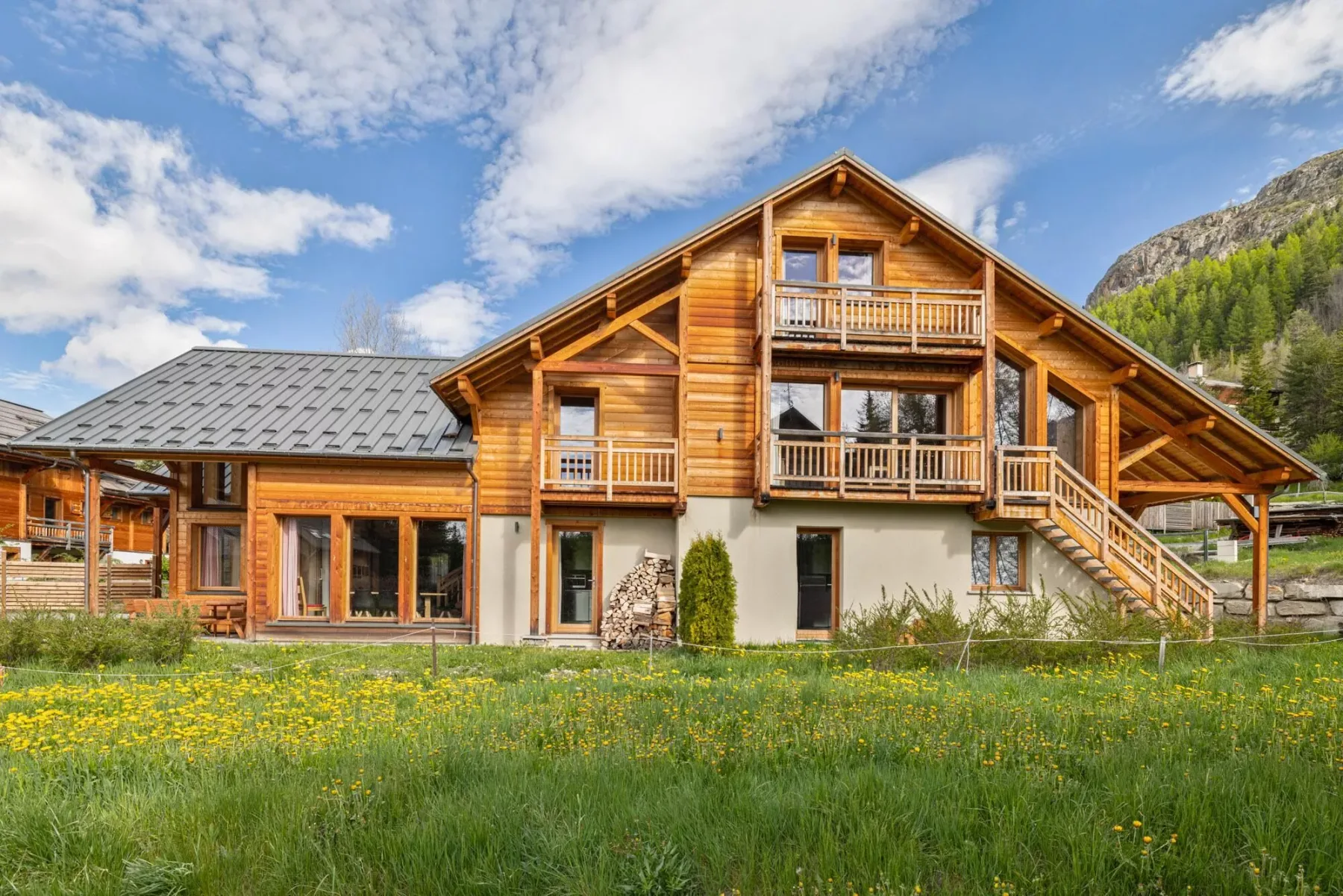 A large wooden chalet stands beside a grassy meadow beneath a partly cloudy sky.