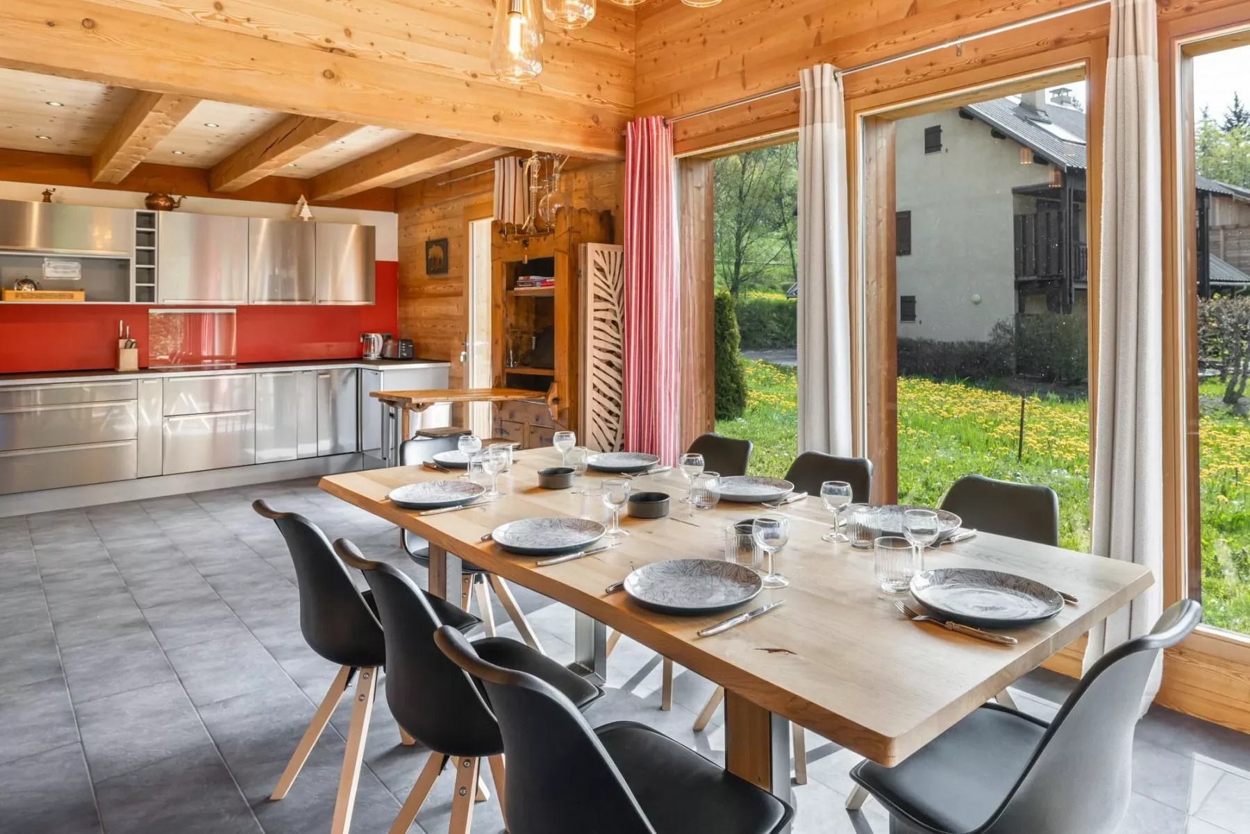 A dining area with a long table beside large windows, next to an open kitchen.