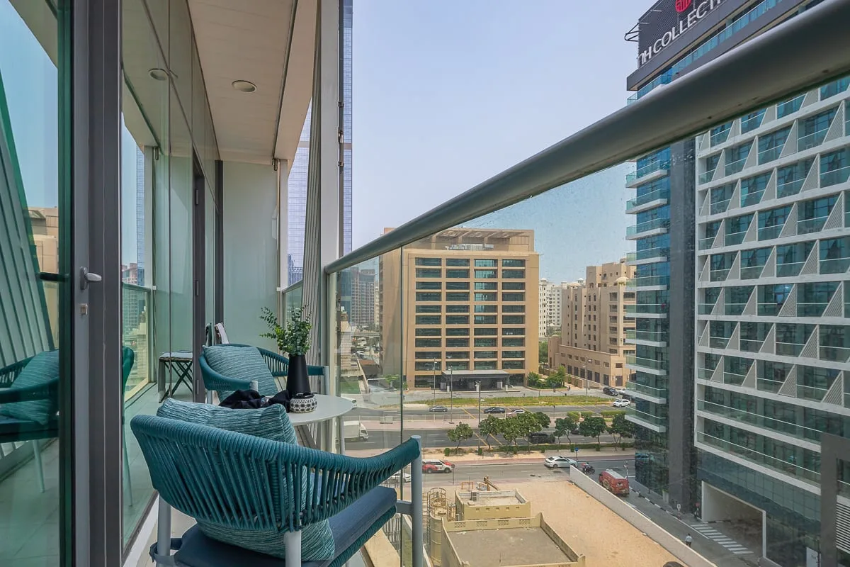 A balcony seating area overlooks nearby modern buildings and a tree-lined street.