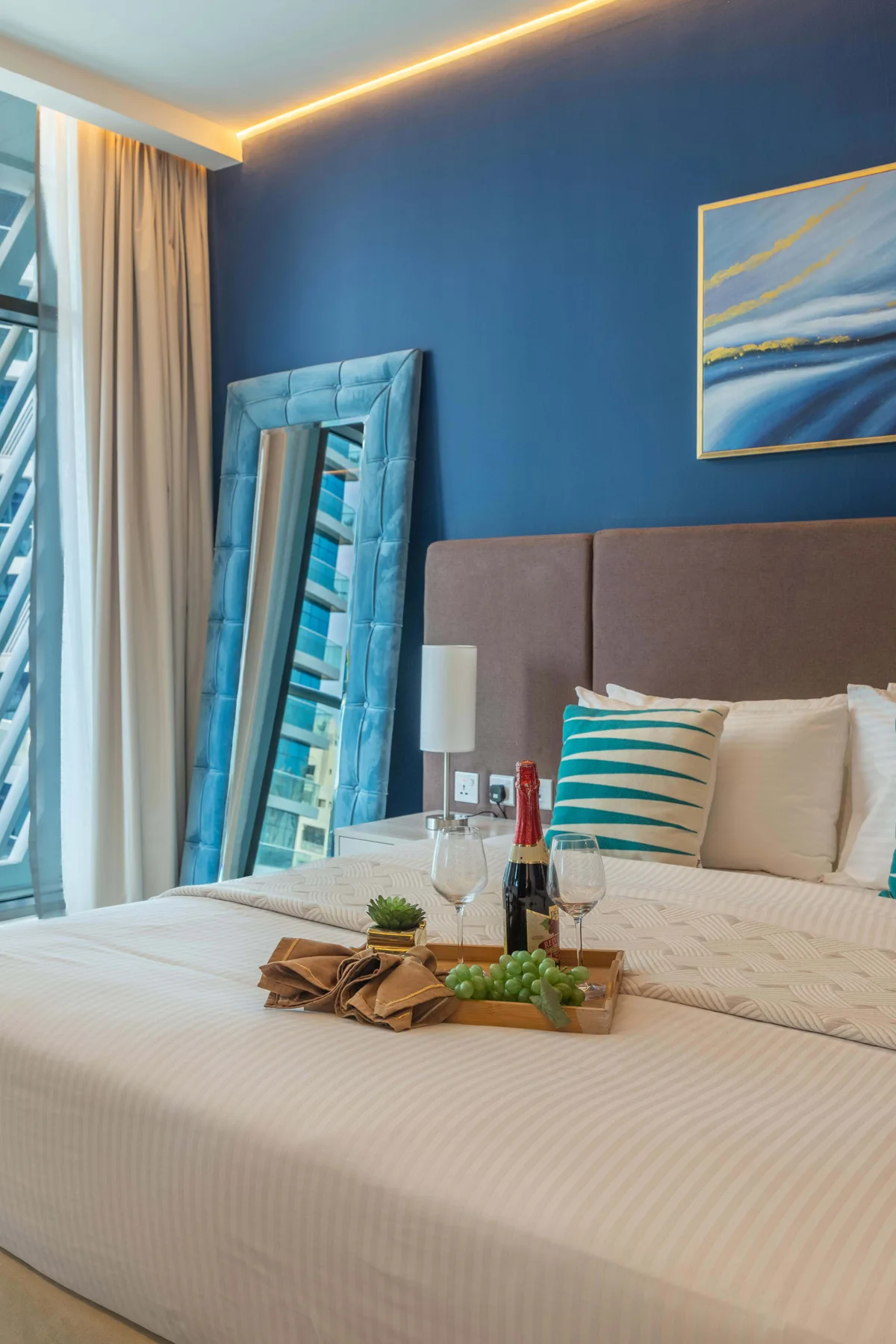 A bedroom with a neatly made bed, blue accent wall, and large leaning mirror.