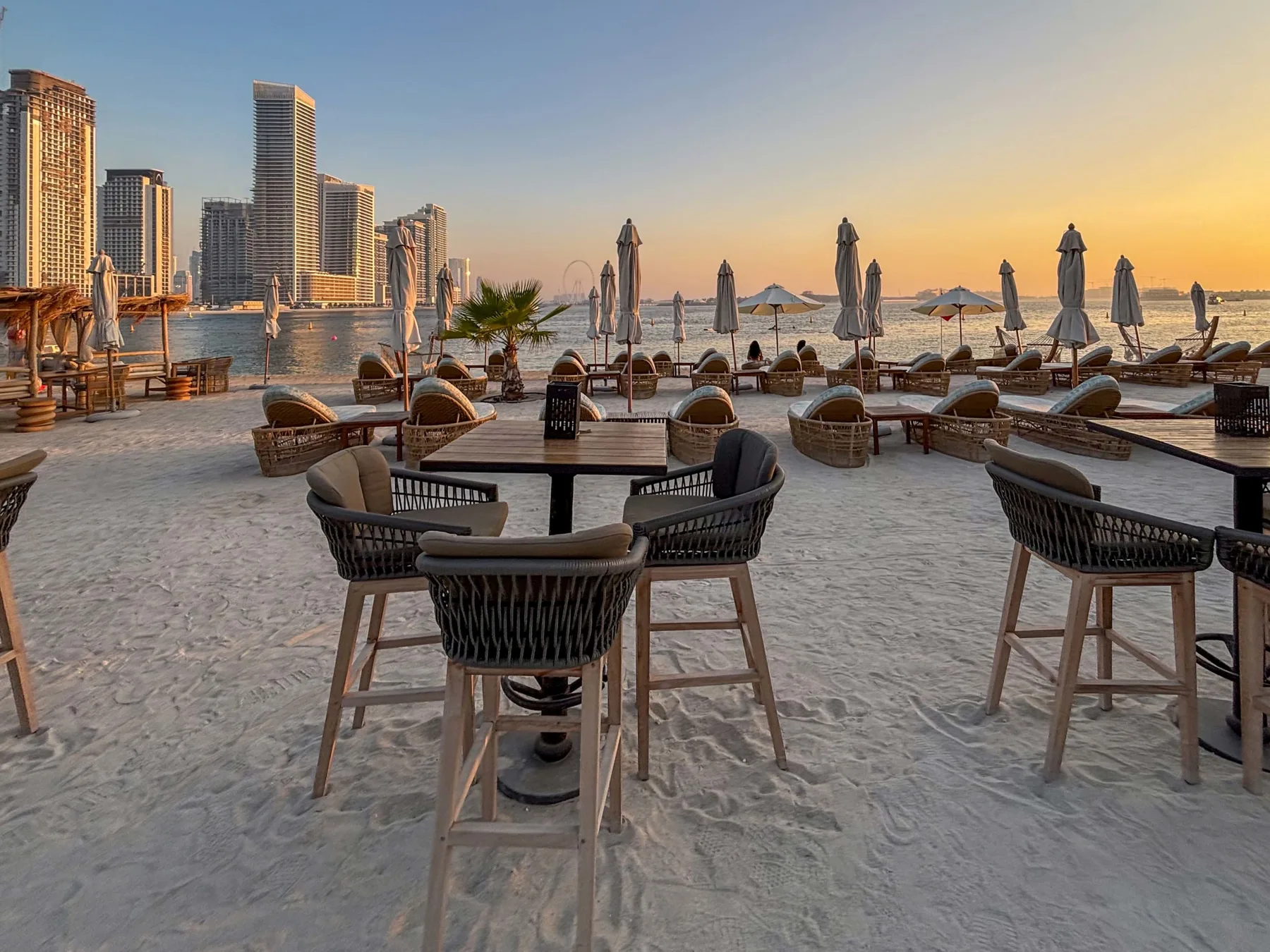 A beachfront dining area with tables, woven seating, and umbrellas beside the water.