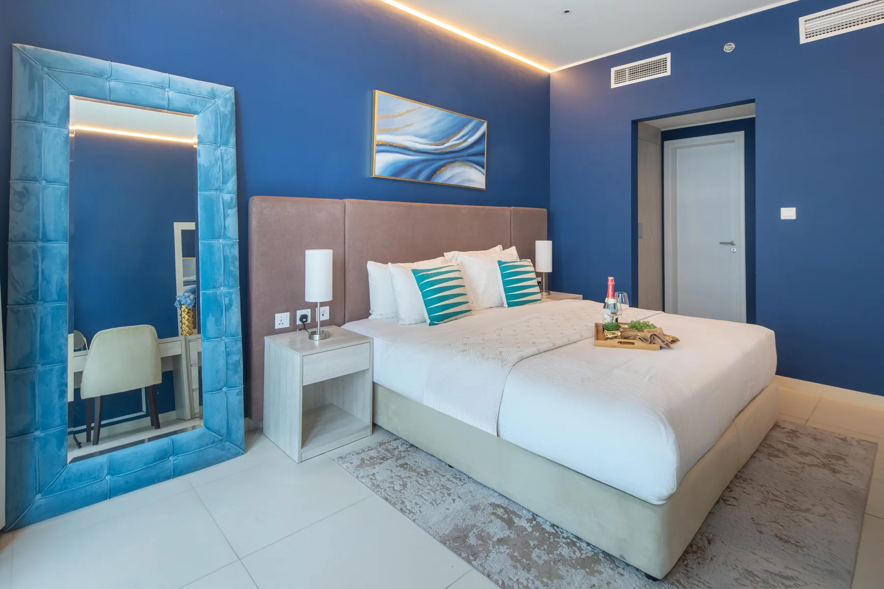 A bedroom with a neatly made bed, blue walls, and a large mirror.