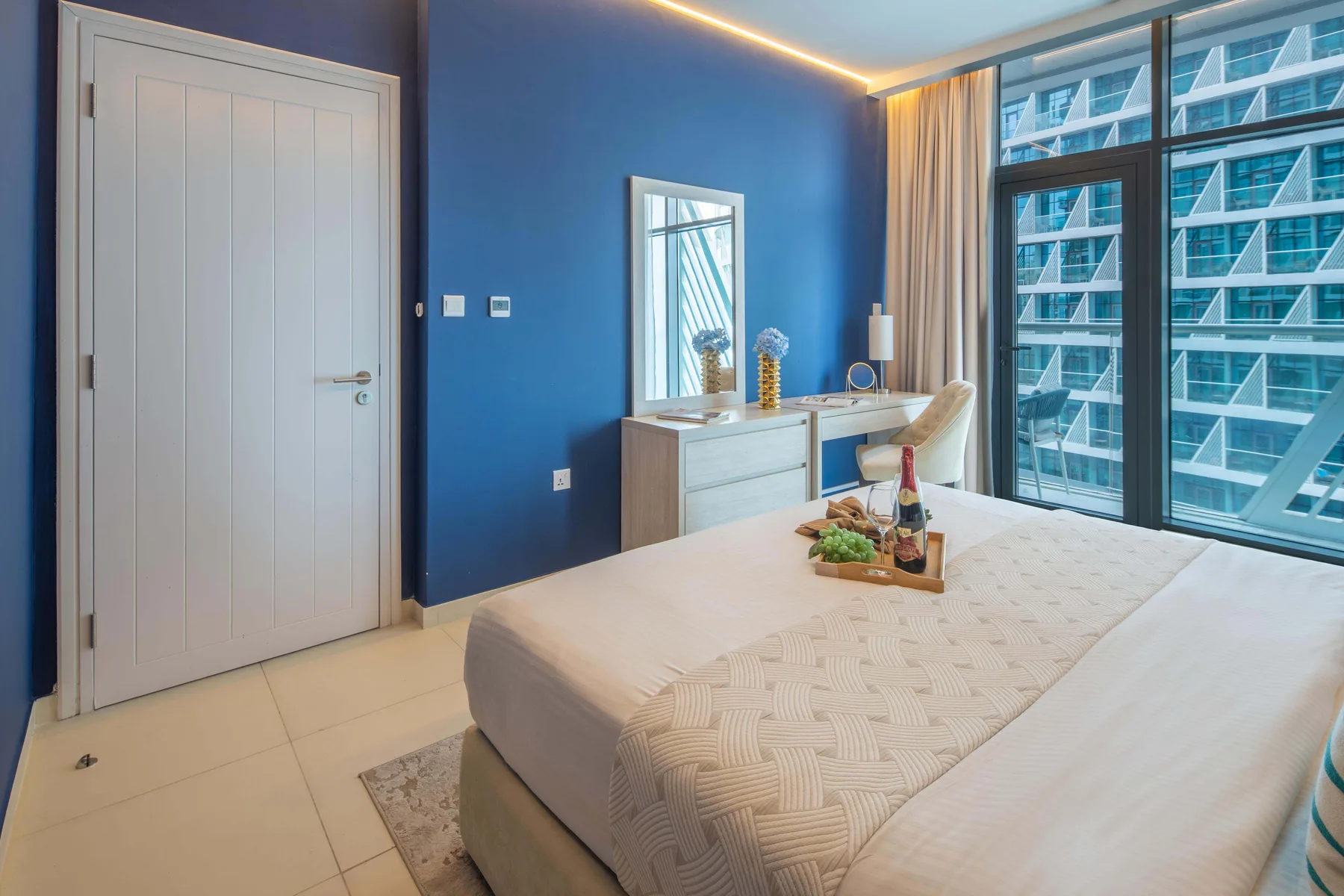 A bedroom with a bed beside large windows overlooking nearby buildings.