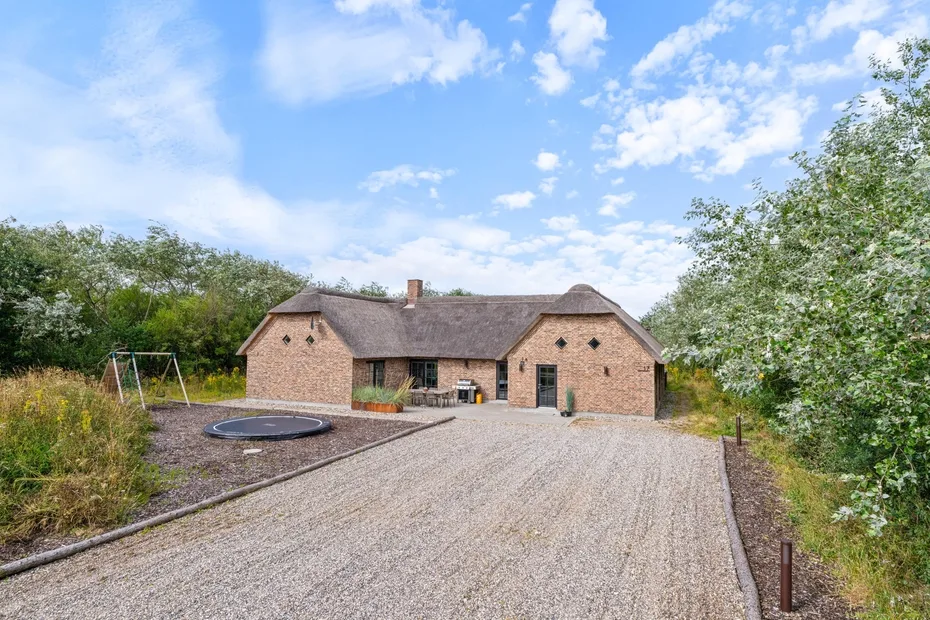 A brick house stands beside a gravel driveway, with a trampoline and swing set nearby.