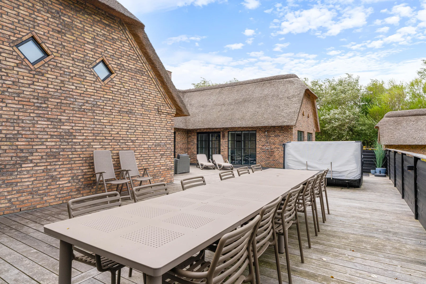 An outdoor dining terrace with a long table and seating beside a brick building.