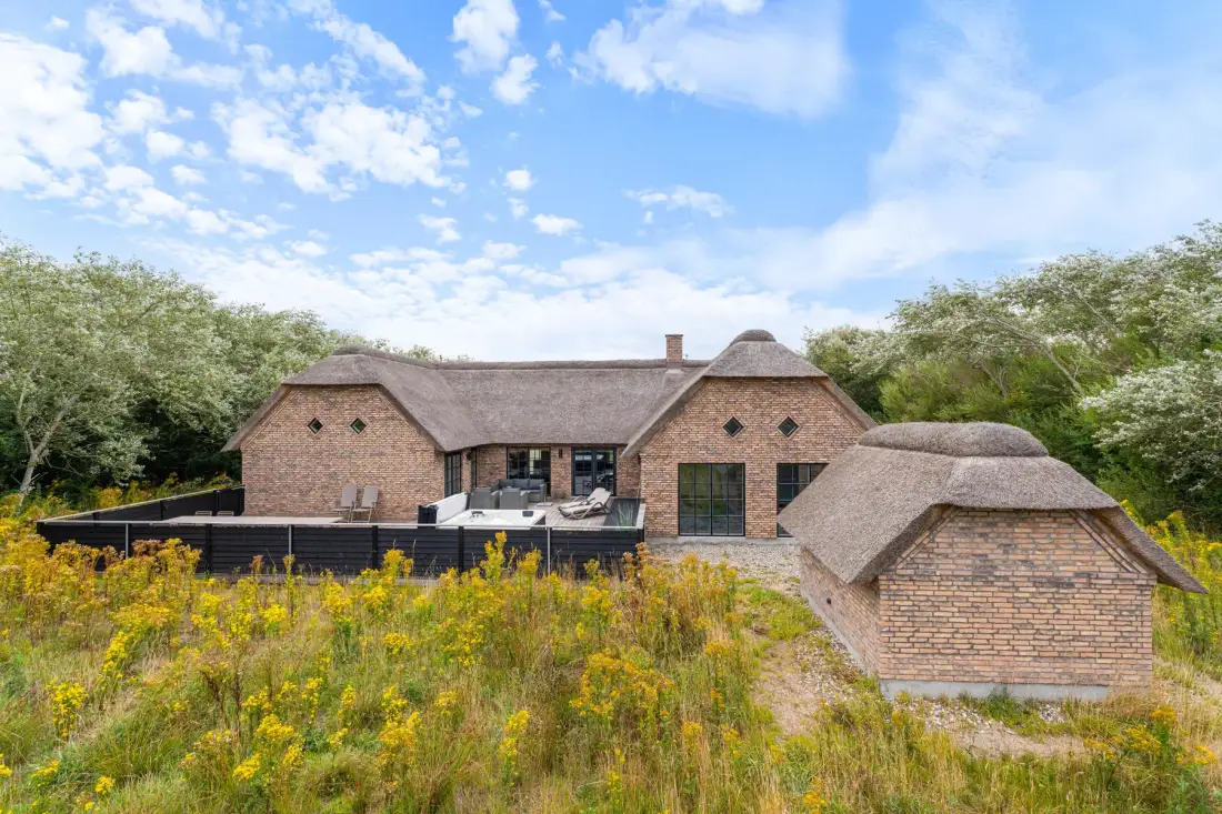 A brick house with thatched roofs stands among trees and wildflowers.