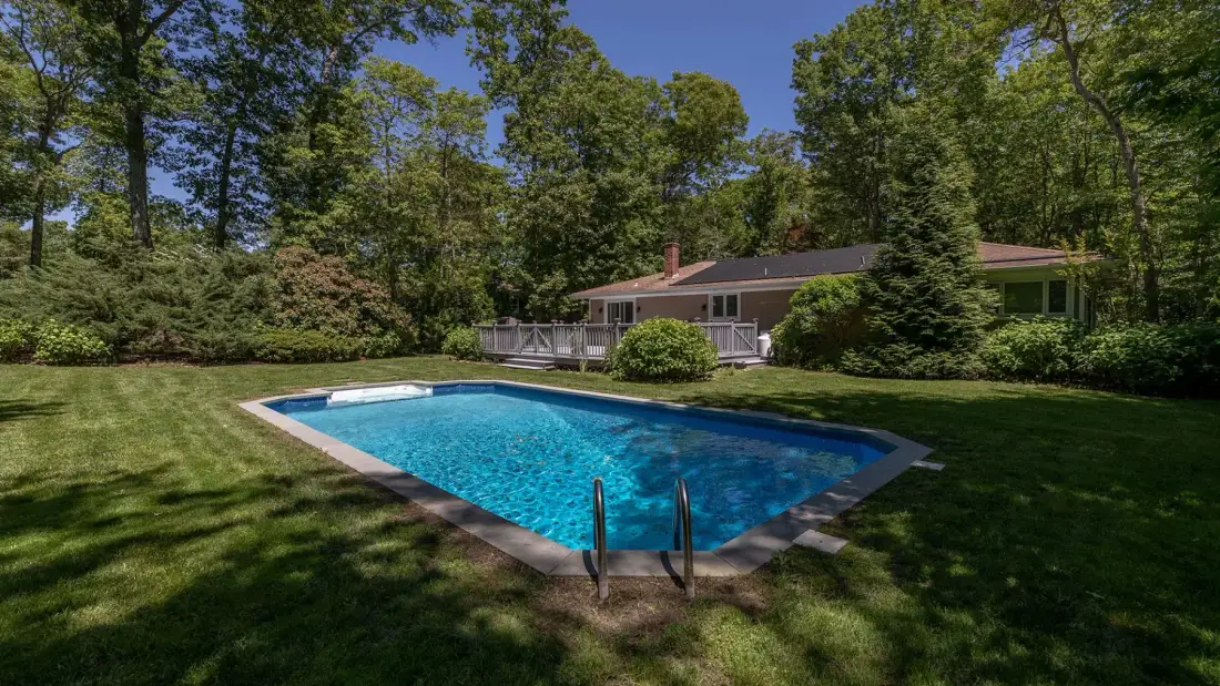 A swimming pool sits in a grassy yard with a house and trees behind it.