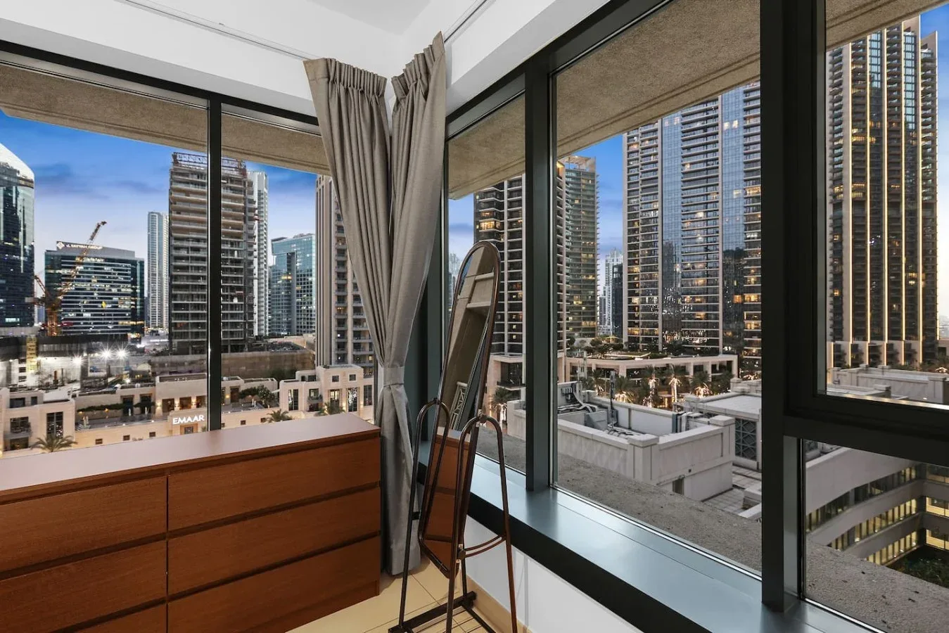 Una habitación con grandes ventanales da a un horizonte urbano, junto a una cómoda y cortinas.