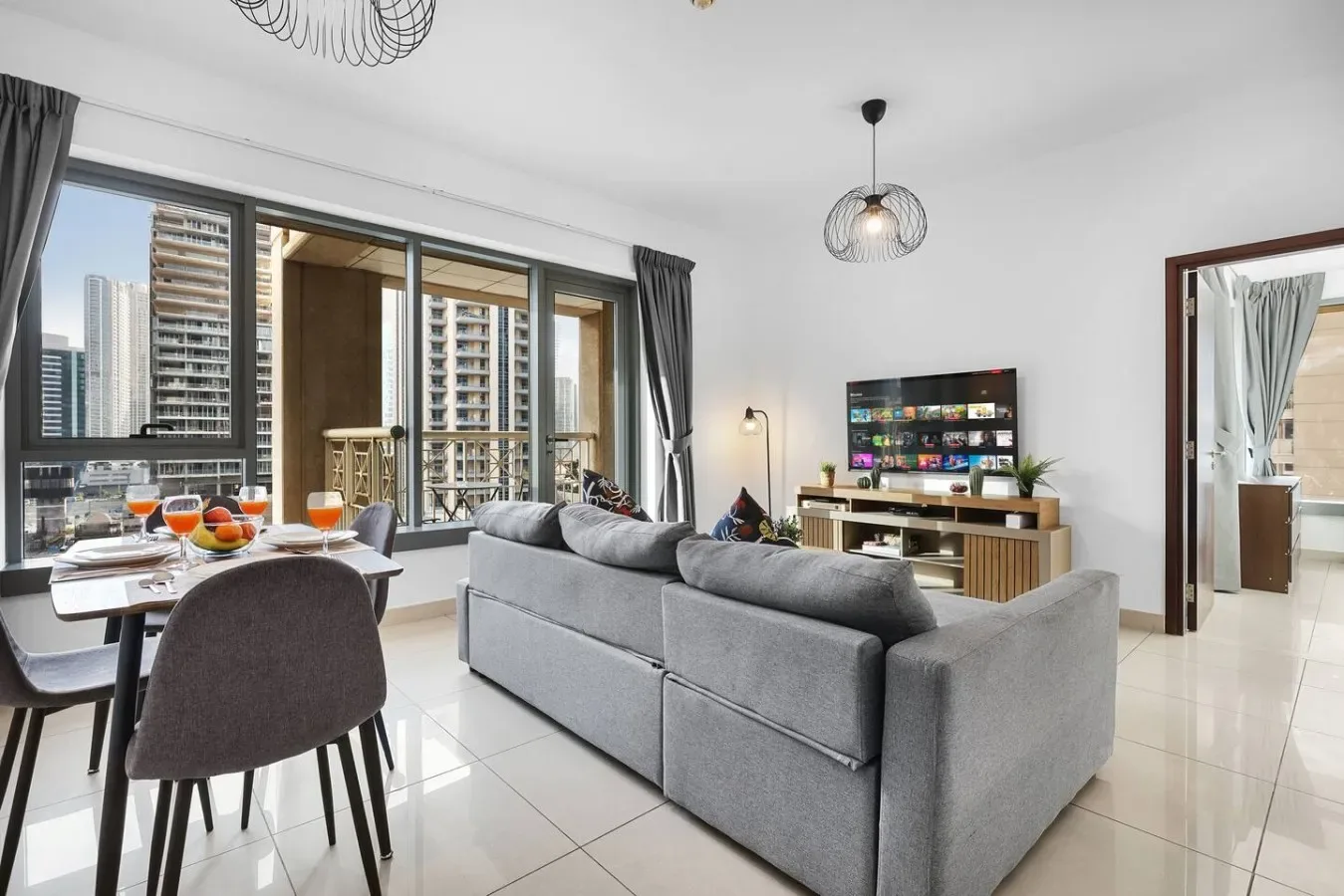 Una sala de estar con sofá gris, televisor y comedor junto a grandes ventanas.