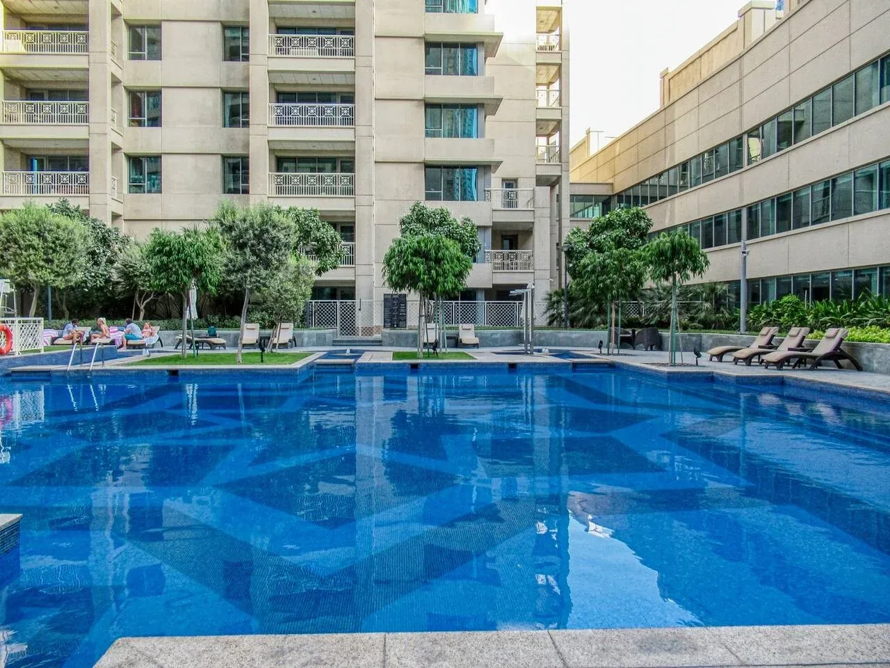 Una piscina al aire libre junto a edificios altos, con árboles y asientos de descanso en el borde.
