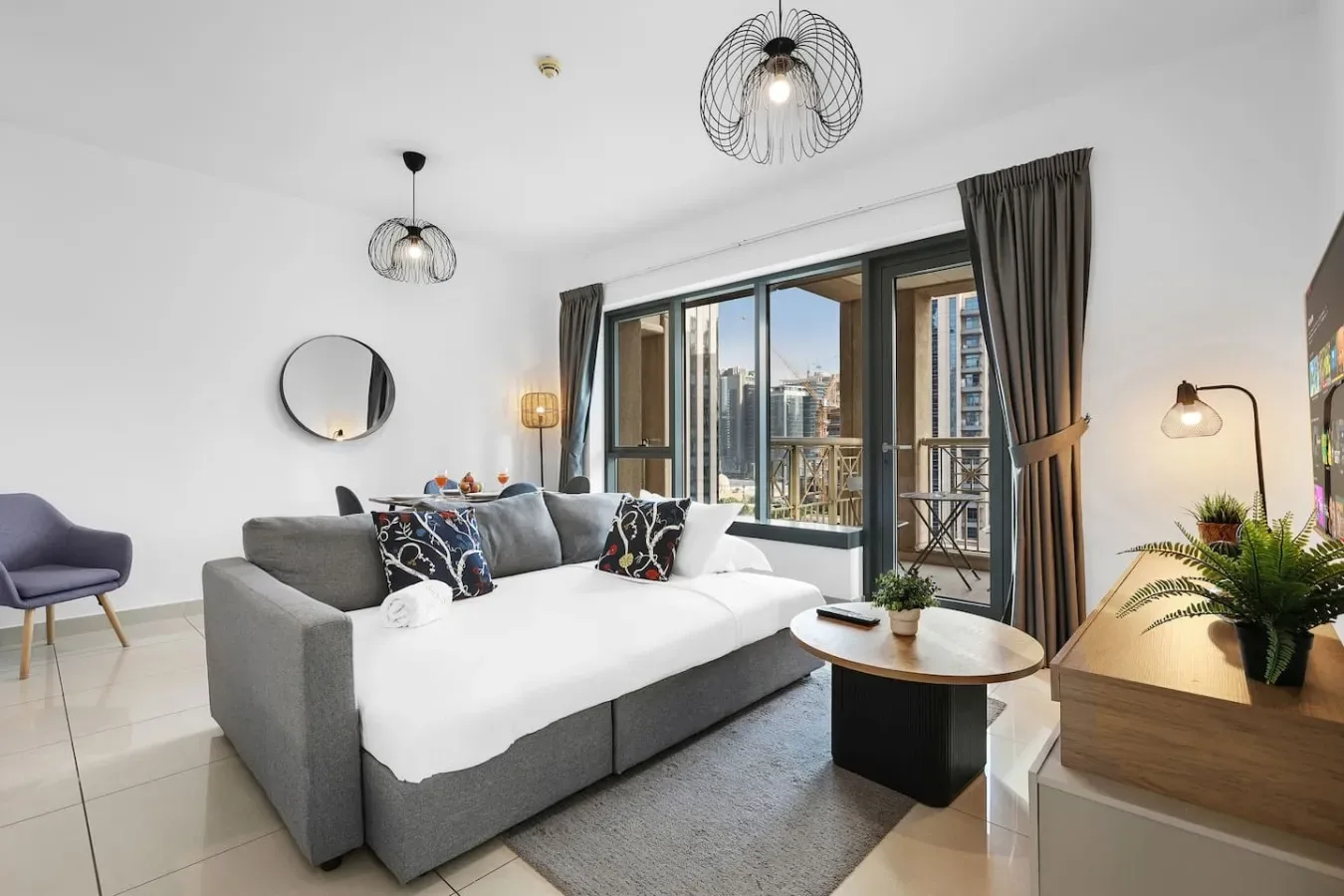 Una sala luminosa con un sofá gris frente a grandes ventanas y vistas de la ciudad.