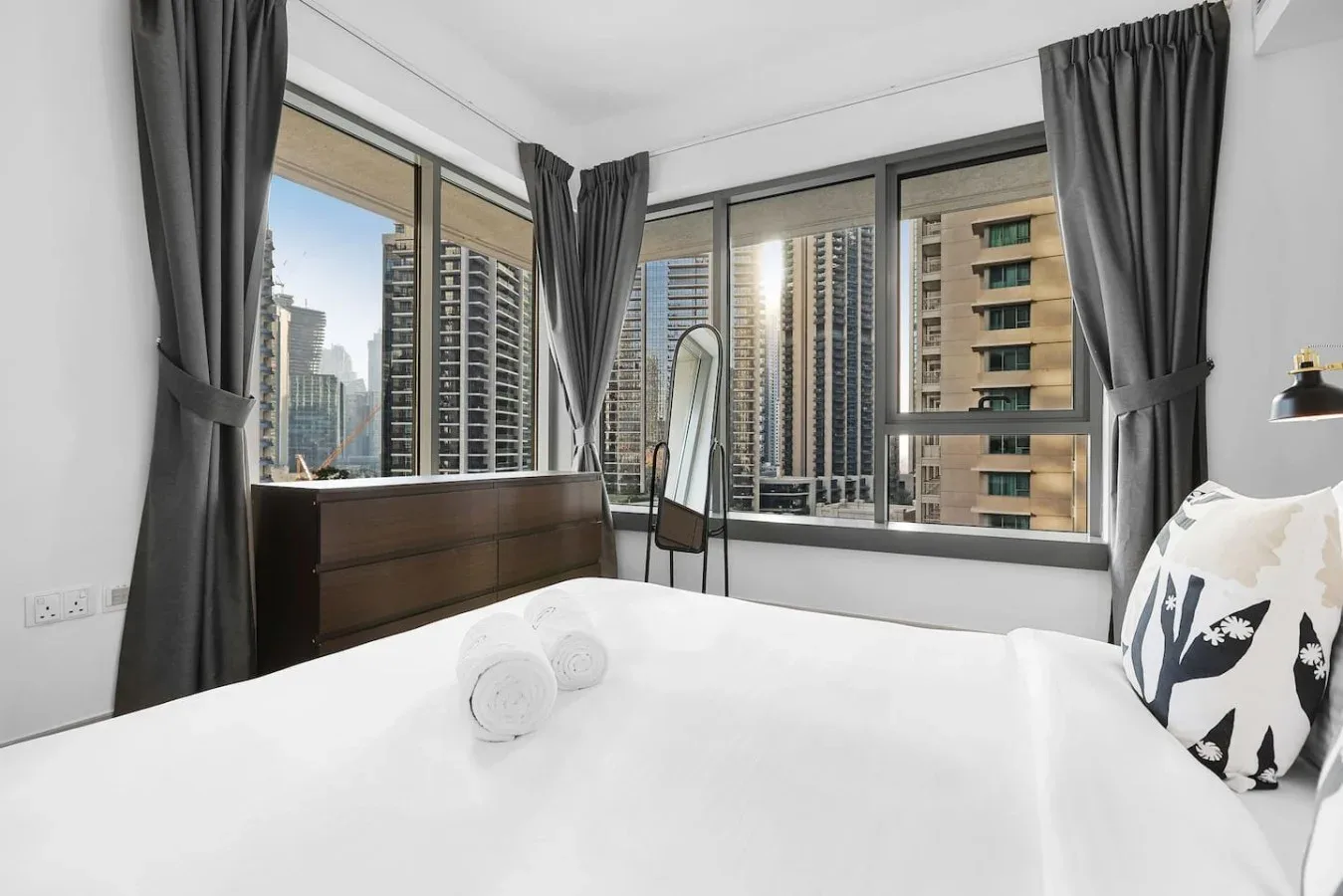 Dormitorio con una cama junto a grandes ventanas con vistas a edificios cercanos.