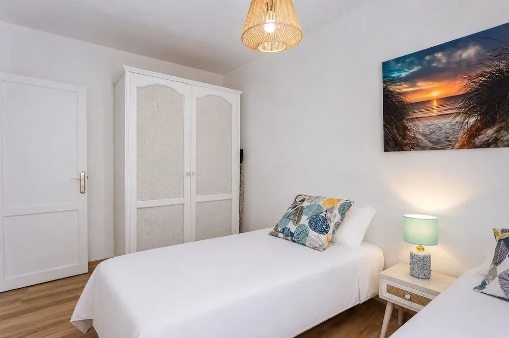 A bright bedroom with white bedding, a wardrobe, and a framed beach picture.