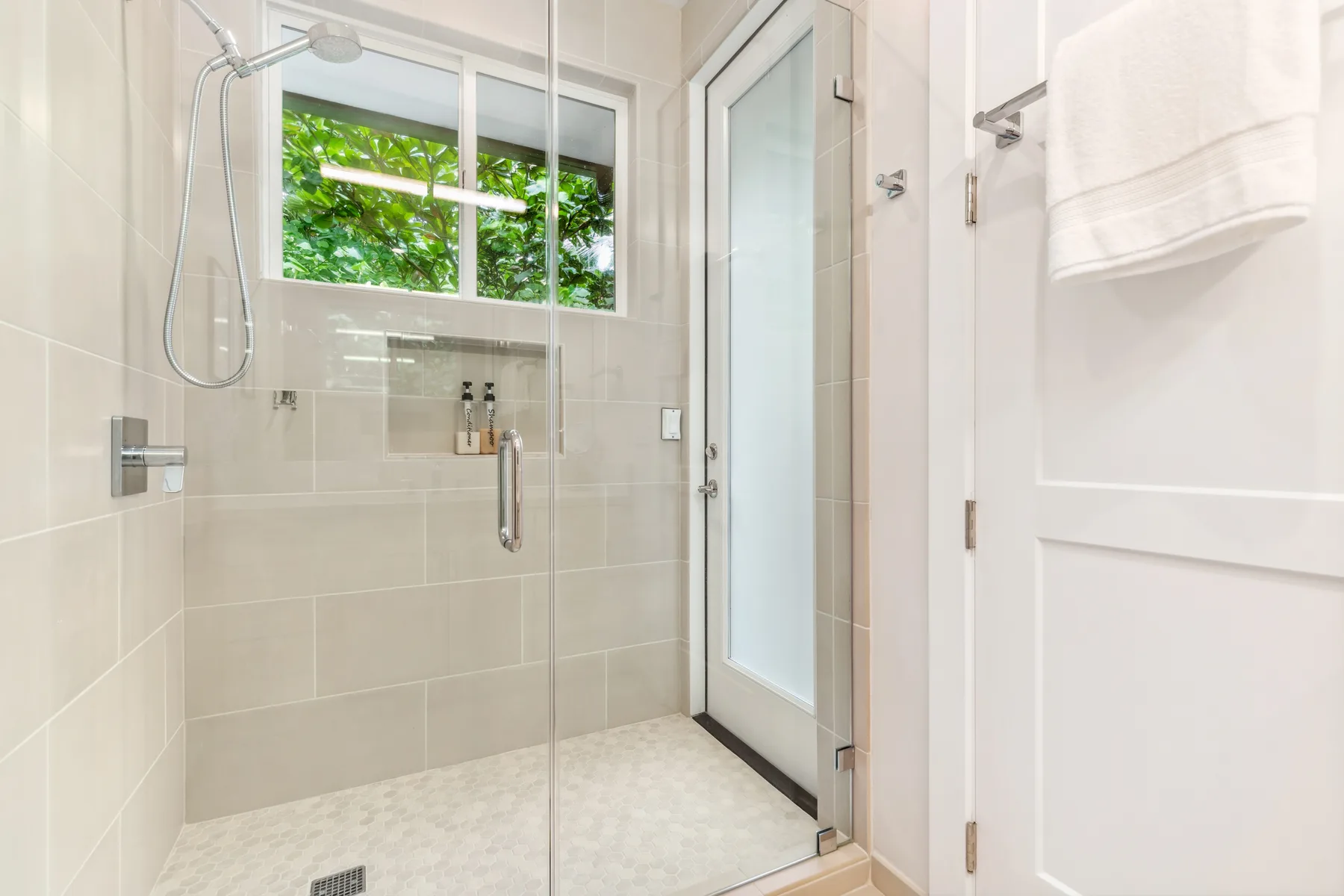 Un baño con azulejos, una ducha de vidrio y una ventana con vista a la vegetación.