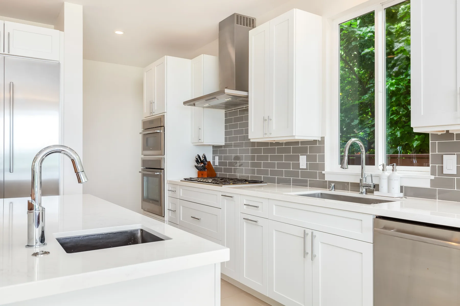 Una cocina luminosa con gabinetes blancos, azulejos grises y una gran ventana con vegetación al fondo.
