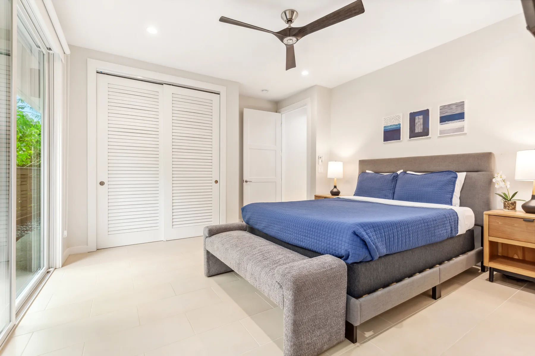 Un dormitorio luminoso con una cama azul, un banco tapizado gris y un ventilador de techo.