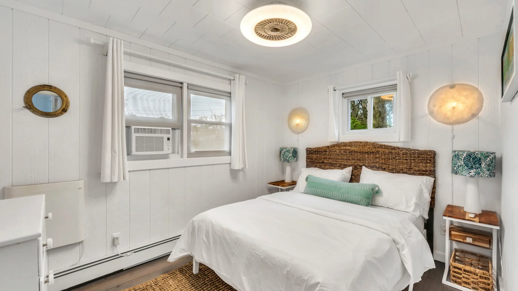A bright bedroom with a neatly made bed and windows along two walls.