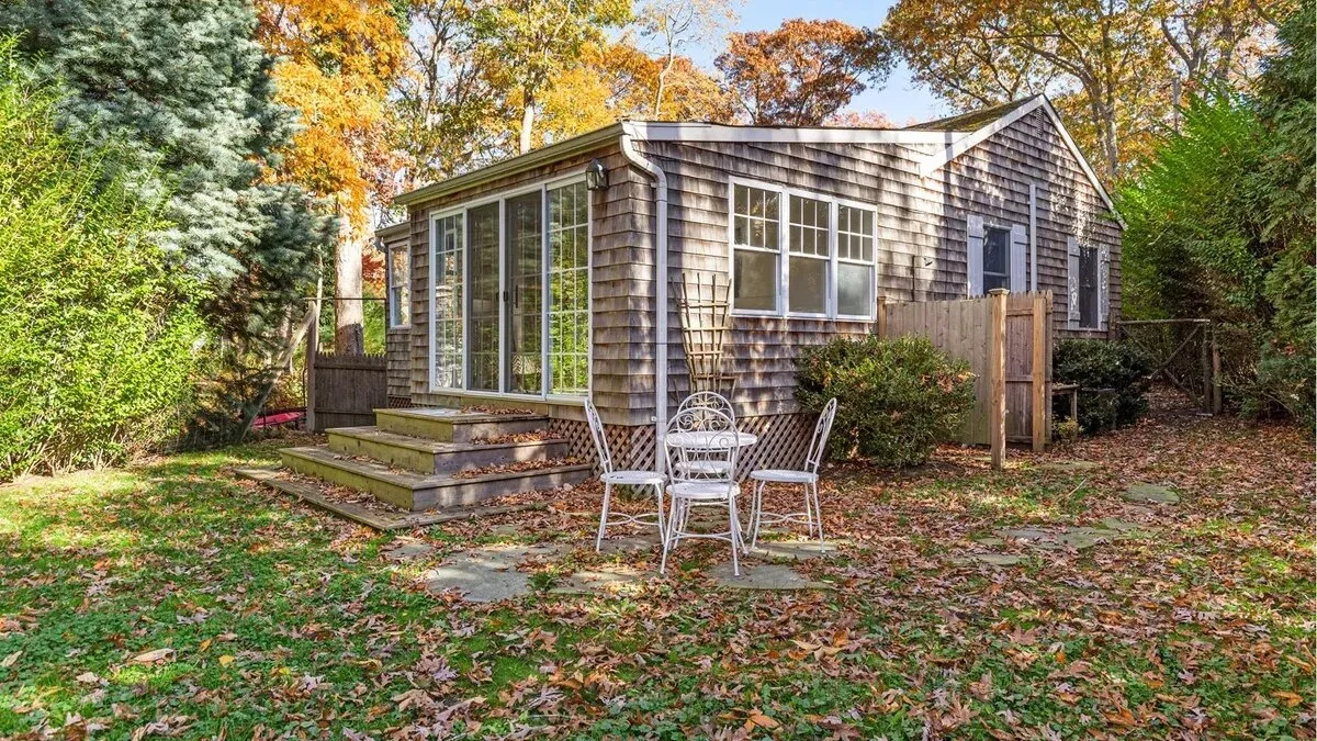 A small cottage with a raised deck and outdoor seating sits among autumn trees.