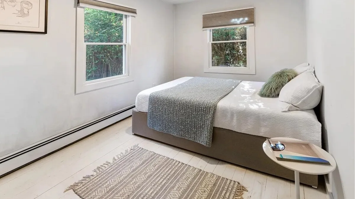 A bright bedroom with a bed, woven rug, and two windows.