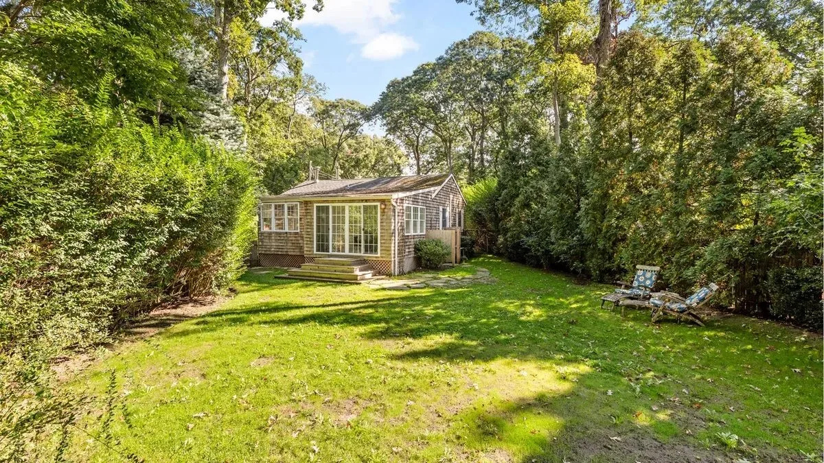 A small garden cottage sits at the edge of a sunny lawn surrounded by trees.