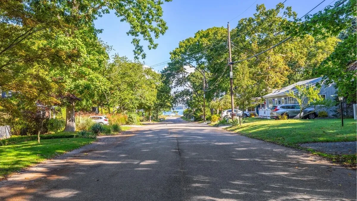 A tree-lined residential street with houses, parked vehicles, and overhead utility wires.