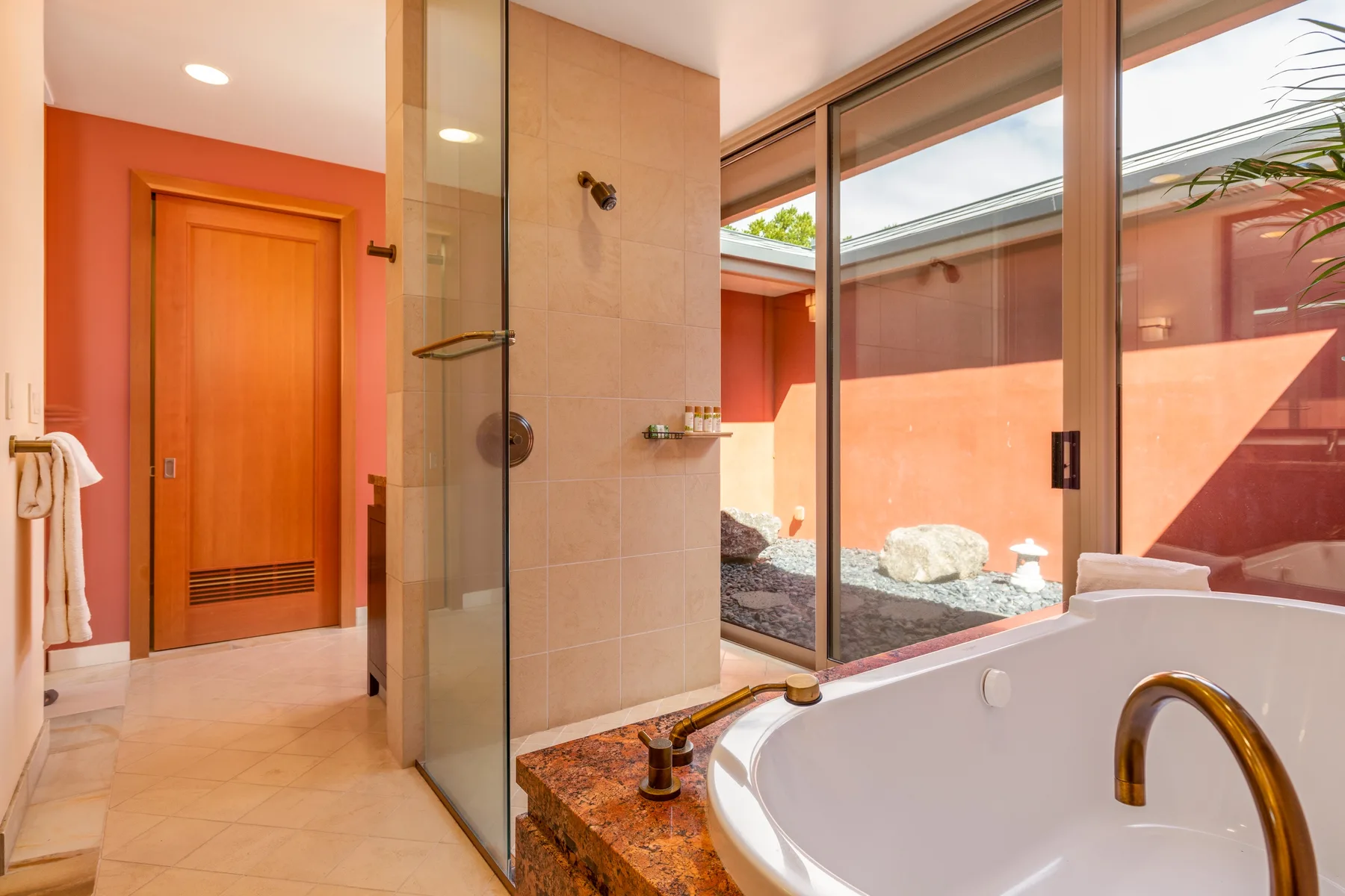 Un baño con ducha de vidrio junto a una bañera y ventanas con vistas a un patio amurallado.