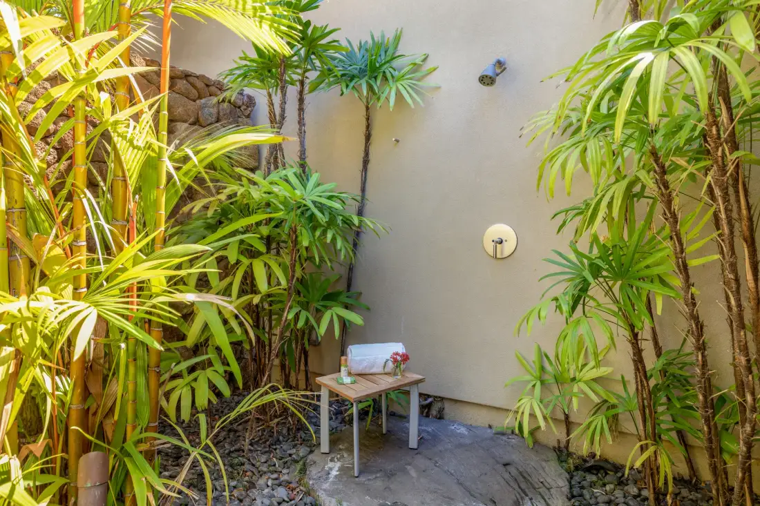 Una ducha exterior rodeada de plantas, con una mesa pequeña junto a la pared.