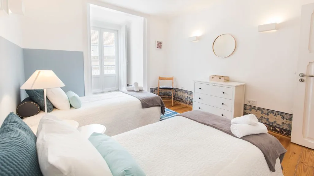Un dormitorio luminoso con dos camas, una cómoda blanca y un espejo redondo en la pared.
