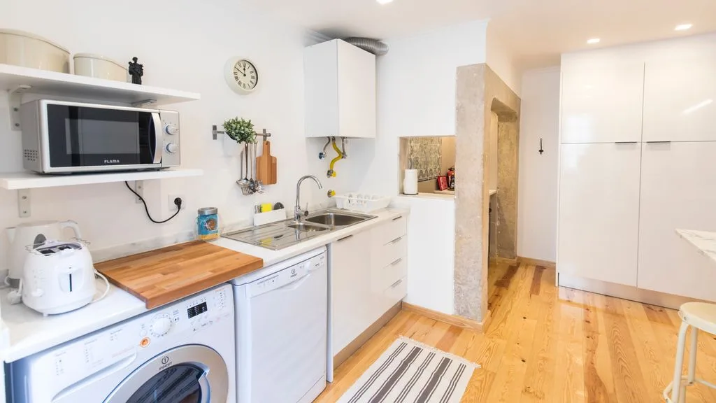 Una cocina luminosa con armarios blancos, electrodomésticos y suelo de madera.