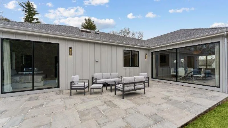 A paved patio with outdoor seating sits beside a house with large glass doors.
